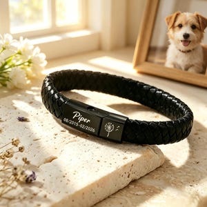 Puede incluir: Pulsera de cuero trenzado negro con cierre negro. El cierre está grabado con el nombre "Piper", fechas y un diseño de diente de león. La pulsera se exhibe sobre una superficie texturizada, con un retrato de perro enmarcado en el fondo.