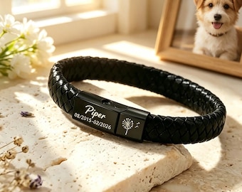 Op de afbeelding: Zwart gevlochten leren armband met een zwarte sluiting. De sluiting is gegraveerd met de naam "Piper", data en een paardenbloemontwerp. De armband wordt getoond op een getextureerd oppervlak, met een ingelijst hondenportret op de achtergrond.