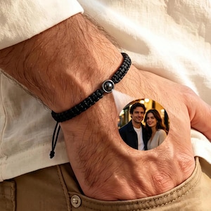 Könnte beinhalten: Ein schwarzes Armband aus geflochtener Kordel mit einem runden Anhänger in der Mitte. Der Anhänger enthält eine kleine, kreisförmige, farbige Fotoprojektion eines Mannes und einer Frau, die zusammenstehen und durch die dunkle Metallperle sichtbar ist.