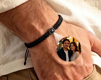 Puede incluir: Una pulsera de cordón trenzado negro con un dije circular central. El dije contiene una pequeña proyección fotográfica circular a todo color de un hombre y una mujer de pie juntos, visible a través del centro de la cuenta metálica oscura.