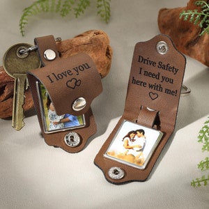 以下が含まれることがあります： 銀色の金属フレームが付いた、茶色の革製のフォトキーホルダーが2つ。1つのキーホルダーには「愛してる」という文字とハートが2つ。もう1つのキーホルダーには「安全運転で！あなたがいなきゃダメなんだ！一緒にいたい！」という文字とハートが1つ。どちらのキーホルダーにも、カップルの写真が飾られています。