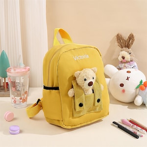 May include: A bright yellow backpack featuring the name Victoria embroidered in white. The front includes a corduroy pocket containing a small, plush beige teddy bear with a brown nose. The bag has yellow straps and a top handle for carrying.