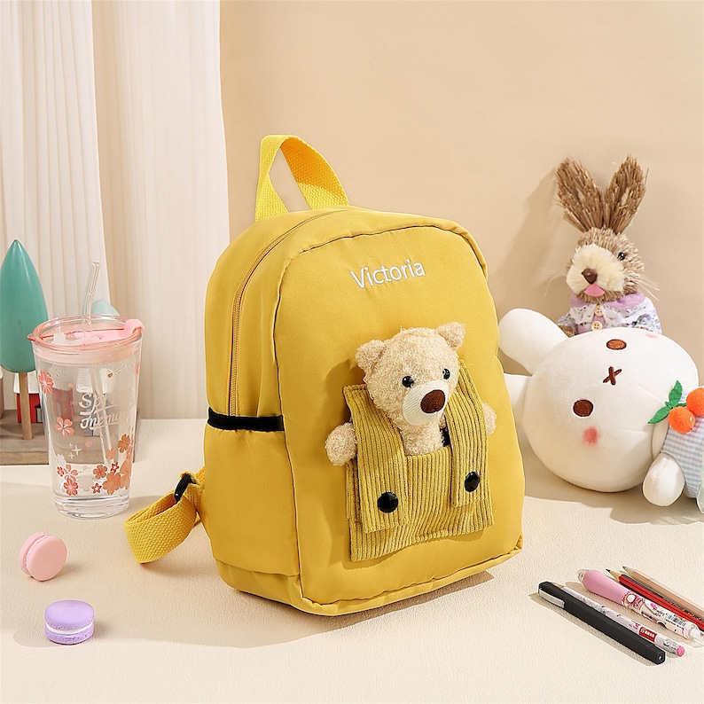 May include: A bright yellow backpack featuring the name Victoria embroidered in white. The front includes a corduroy pocket containing a small, plush beige teddy bear with a brown nose. The bag has yellow straps and a top handle for carrying.