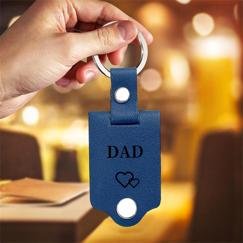 May include: A blue leather keychain with the word "DAD" and two heart symbols. The keychain has a silver metal ring and two white button-like fasteners. The background is blurred, suggesting an indoor setting.