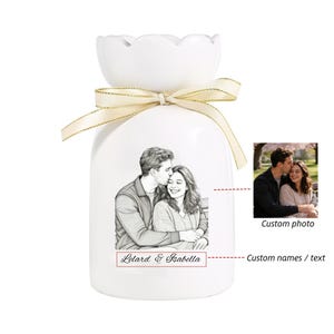 Puede incluir: Un jarr&oacute;n de cer&aacute;mica blanca con una parte superior festoneada y una cinta dorada. El jarr&oacute;n presenta un boceto a l&aacute;piz de una pareja y los nombres "Letard & Isabella". Tambi&eacute;n se incluye una peque&ntilde;a foto de la pareja.
