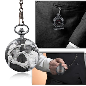 Op de afbeelding: Een zilveren zakhorloge met een zwarte wijzerplaat en een foto van een ouder koppel gegraveerd op de voorkant. Het horloge zit aan een ketting en hangt aan een colbert.