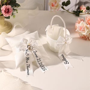 May include: A white satin ring pillow with a large bow and a ribbon with the text "Mr & Mrs Thomas" and "June 21, 2025". A white flower girl basket with a pearl handle and a ribbon with the name "Selena".