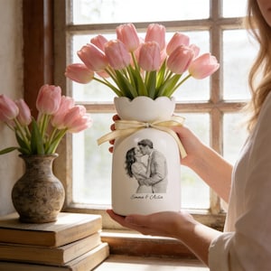 Puede incluir: Un jarr&oacute;n de cer&aacute;mica blanco con borde festoneado, lleno de tulipanes rosas y adornado con una cinta beige. El jarr&oacute;n presenta una imagen en blanco y negro de una pareja bes&aacute;ndose, con el texto "Emma & Aclia" debajo.