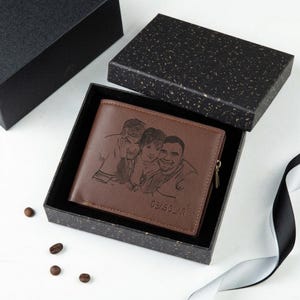 May include: A brown leather wallet with a custom engraved image of three people, presented in a black gift box. The wallet has a zipper and the text "DE/BOLAR" is visible. Coffee beans and a ribbon are also in the image.