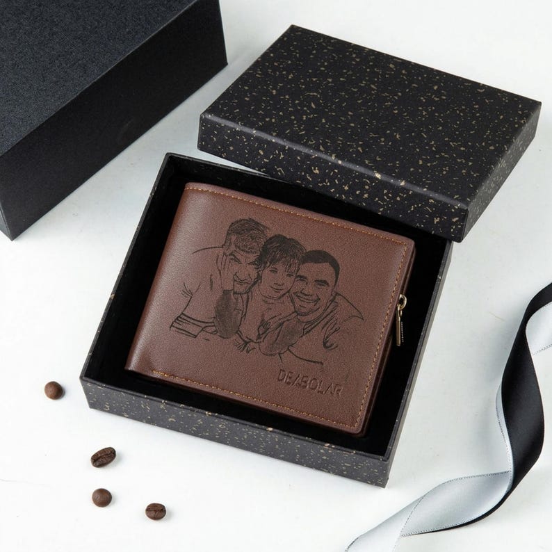 May include: A brown leather wallet with a custom engraved image of three people, presented in a black gift box. The wallet has a zipper and the text "DE/BOLAR" is visible. Coffee beans and a ribbon are also in the image.
