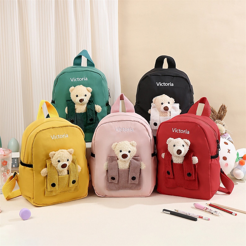 May include: Five small backpacks in yellow, green, black, pink, and red. Each bag features the name Victoria embroidered in white, a top handle, and a plush teddy bear sitting in a front corduroy pocket with two black buttons on the flap.