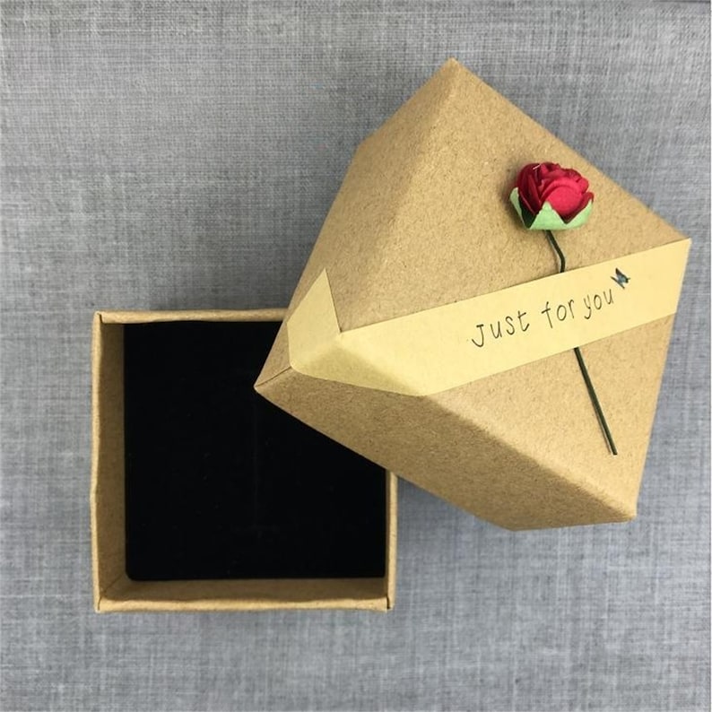 May include: A small, square, brown cardboard gift box with a black velvet interior. The box is open and the lid has a brown paper label with the text "Just for you" and a red paper flower.