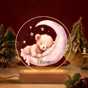 May include: A night light featuring a sleeping teddy bear on a crescent moon, with the name "Olivia" on the moon. The design is set within a clear, circular frame, atop a wooden base. The light emits a warm glow.