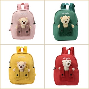 May include: A collection of four small backpacks in pink, green, yellow, and red. Each bag features the name Victoria embroidered in white, adjustable straps, and a plush teddy bear tucked into a corduroy front pocket with two black buttons.