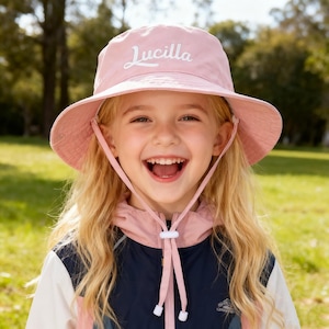 May include: A light pink bucket hat with the name "Lucilla" embroidered in white script. The hat has a wide brim and chin straps. The person in the image is wearing a pink and navy blue jacket.