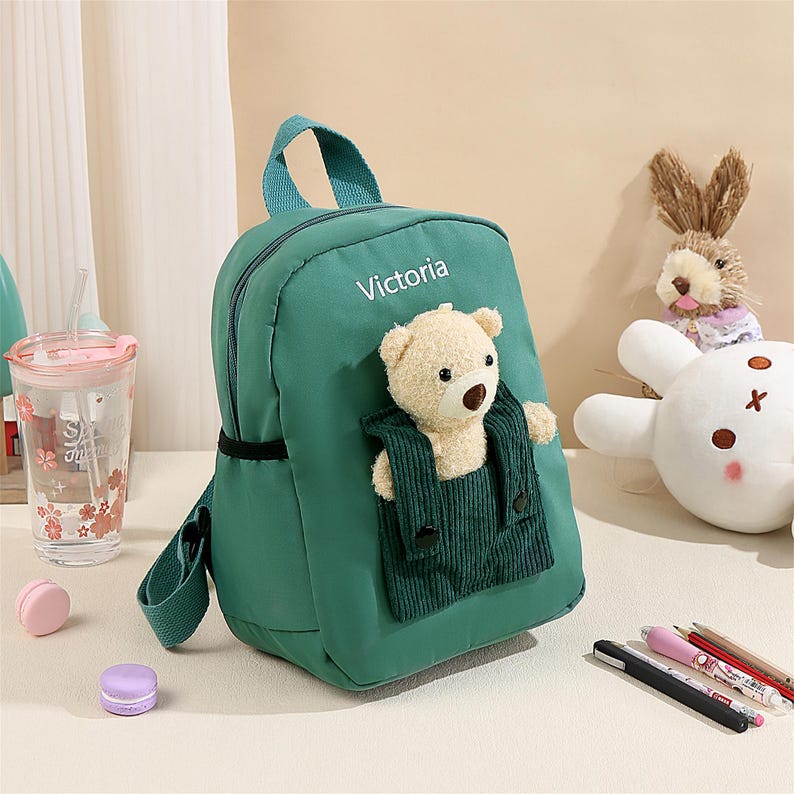 May include: A dark green backpack featuring the name Victoria printed in white text. The bag includes a front pocket made of dark green corduroy fabric, which contains a small, light beige plush teddy bear with a brown nose and black eyes.