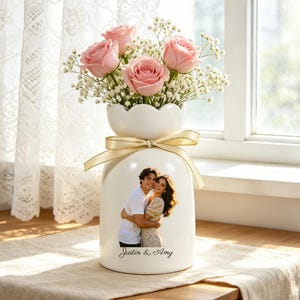 Puede incluir: Un jarr&oacute;n de cer&aacute;mica blanco con la parte superior festoneada, lleno de rosas rosas y paniculata. Una foto de una pareja est&aacute; en el jarr&oacute;n, con el texto "Justin & Amy" debajo. Una cinta color crema est&aacute; atada alrededor del jarr&oacute;n.