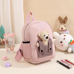 May include: A light pink backpack featuring the name Victoria embroidered in white thread. The front includes a brown corduroy pocket containing a plush beige teddy bear with a dark nose. The bag has matching pink straps and a top handle.