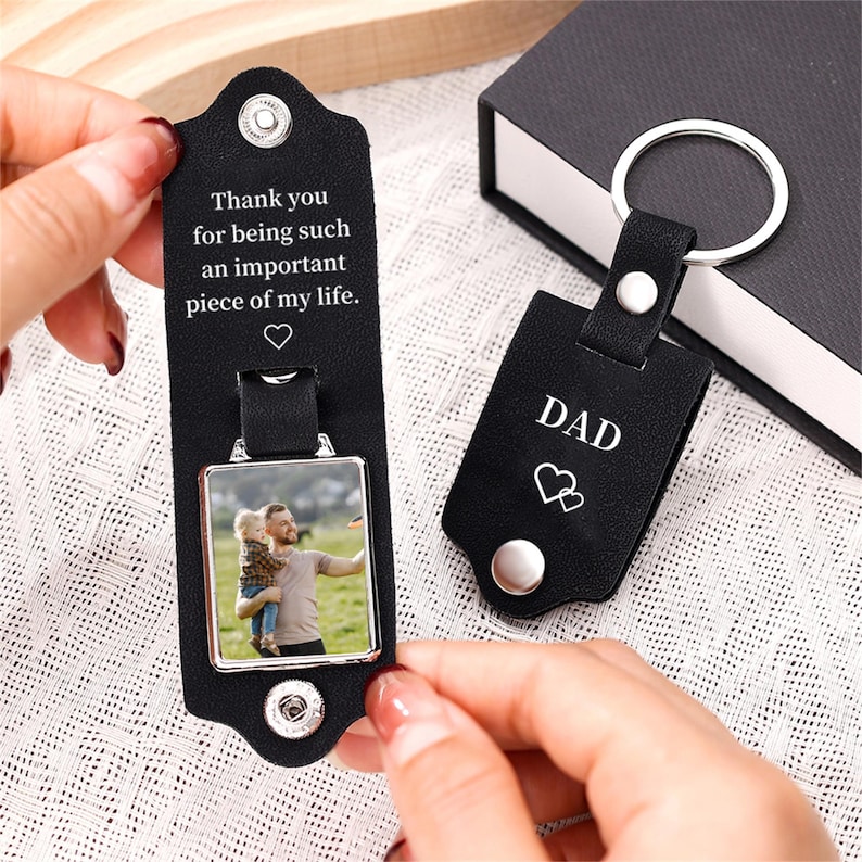 May include: Two black leather keyrings. One keyring has a photo and the text "Thank you for being such an important piece of my life." The other keyring has the word "DAD" and two hearts. Both keyrings have silver hardware.