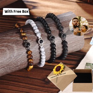 May include: Four beaded bracelets with metal accents. The bracelets are made of black lava rock, white marble, tiger's eye, and black onyx beads. The bracelets are displayed on a wooden surface with a small gift box and a sunflower.