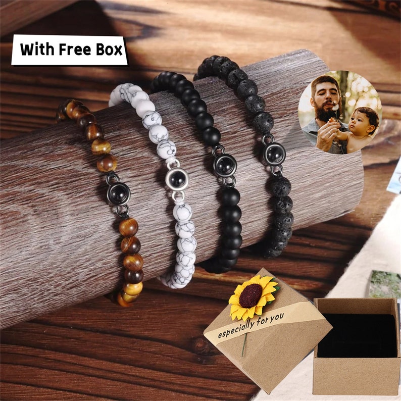 May include: Four beaded bracelets with metal accents. The bracelets are made of black lava rock, white marble, tiger's eye, and black onyx beads. The bracelets are displayed on a wooden surface with a small gift box and a sunflower.
