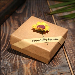 May include: A square, brown gift box with a yellow ribbon that reads "especially for you." A yellow and purple sunflower is attached to the top of the box. The box is on a dark wooden surface.