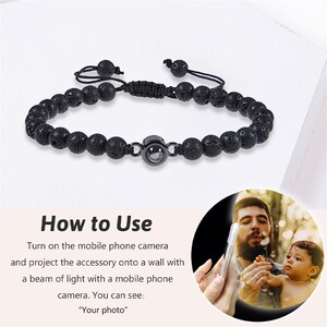 May include: A black lava rock beaded bracelet with a small round charm that projects an image when held up to a phone camera. The charm has a silver ring around it. "How to Use" is written in white text on a pink background with instructions on how to use the bracelet.