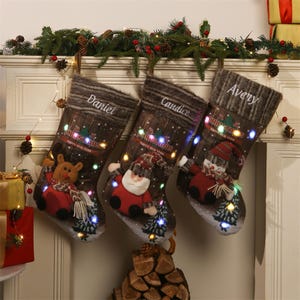 Peut inclure: Trois bas de Noël décoratifs sont suspendus à une cheminée blanche. Chaque bas présente un personnage différent : un renne, le Père Noël et un bonhomme de neige. Les bas sont éclairés par de petites lumières colorées et portent les noms "Daniel", "Candice" et "Avery".