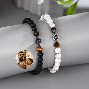 May include: Two beaded bracelets with a photo charm. One bracelet is black with brown beads and the other is white with brown beads. Both bracelets have silver accents.