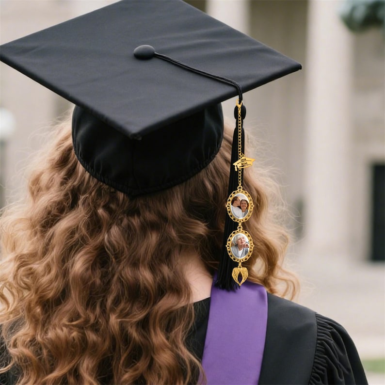 Pode incluir: Um chap&eacute;u de formatura preto com uma borla dourada e um pingente decorativo. O pingente apresenta duas molduras ovais para fotos e um pingente de chap&eacute;u de formatura. O formando est&aacute; usando uma beca preta com uma faixa roxa.