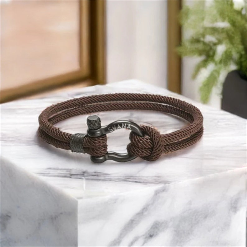 May include: A brown rope bracelet with a black metal clasp. The clasp has the word "SVANA" engraved on it.
