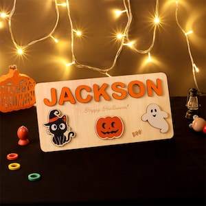 Pode incluir: Um quebra-cabeça de Halloween de madeira personalizado com o nome "JACKSON" em letras laranja. O quebra-cabeça apresenta um gato preto, uma abóbora, um fantasma e o texto "Feliz Halloween!" e "Boo!"