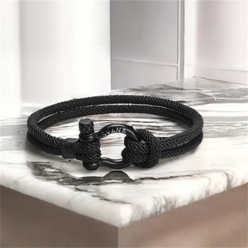 May include: Black rope bracelet with a black metal clasp. The clasp has the word "SVANA" engraved on it.
