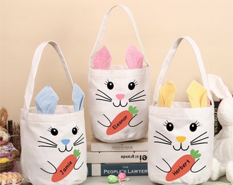Puede incluir: Tres cestas de Pascua de lona blanca con caras de conejo y orejas caídas. Cada cesta tiene una zanahoria y un nombre: Jamie, Eleanor y Herbert.