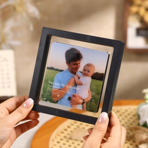 May include: A black square photo frame holding a picture of a man and a baby. The image has the text "You Are My Sunshine" and a music player interface. The frame is held by a person.