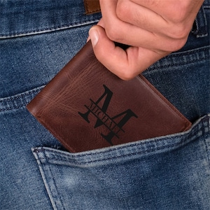 May include: A brown leather wallet with a personalized design is partially visible in a blue jeans pocket. The wallet features a large letter "M" with the name "Michael" underneath, in a black font.