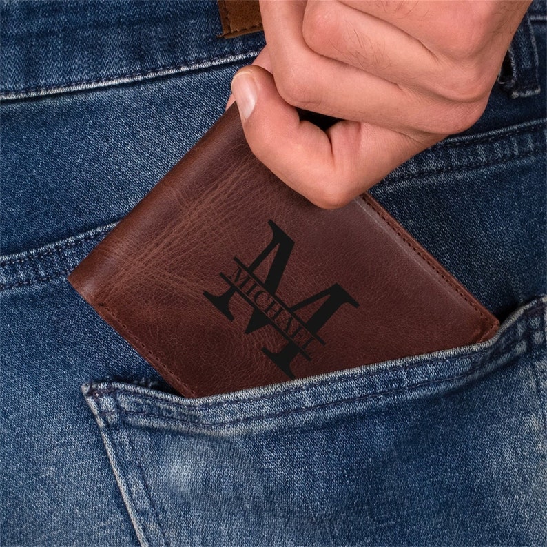 May include: A brown leather wallet with a personalized design is partially visible in a blue jeans pocket. The wallet features a large letter "M" with the name "Michael" underneath, in a black font.