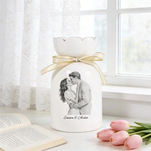 Puede incluir: Un jarr&oacute;n de cer&aacute;mica blanco con una parte superior festoneada y una cinta color crema. El jarr&oacute;n presenta una ilustraci&oacute;n en blanco y negro de una pareja bes&aacute;ndose, con los nombres "Emma & Acilia" debajo. Tulipanes rosas en primer plano.