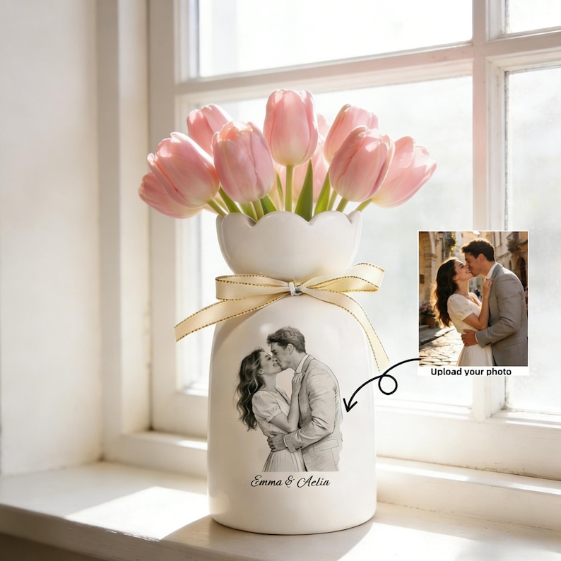 Puede incluir: Un jarr&oacute;n de cer&aacute;mica blanca con borde festoneado contiene tulipanes rosas. El jarr&oacute;n presenta un boceto a l&aacute;piz de una pareja bes&aacute;ndose, con los nombres "Emma & Aelia" debajo. Una cinta est&aacute; atada alrededor del jarr&oacute;n. Se muestra una foto peque&ntilde;a.