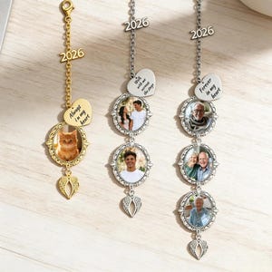 Pode incluir: Tr&ecirc;s amuletos de carro com fotos personalizadas. Um amuleto dourado apresenta uma foto de gato e o texto "Always in my heart". Dois amuletos prateados exibem fotos de pessoas e o texto "Walk with me across the stage" e "Forever in my heart". Cada amuleto inclui o ano "2026".
