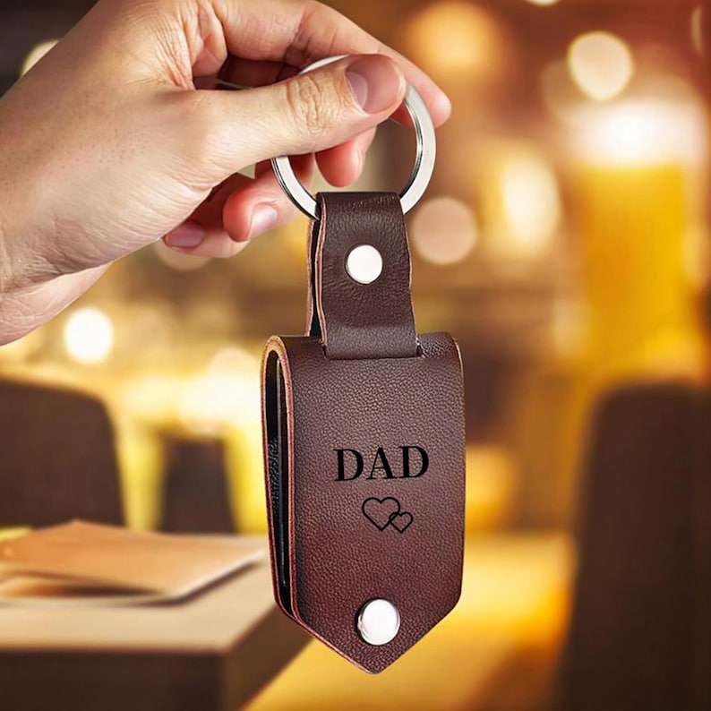 May include: A brown leather keychain with a silver key ring. The keychain features the word "DAD" and two hearts. The keychain is held in a hand.