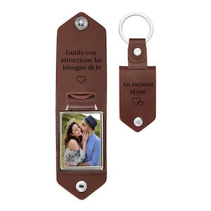 May include: A brown leather keychain and photo holder set. The photo holder has a picture of a couple and the text "Guida con attenzione ho bisogno di te" with a heart symbol. The keychain has the text "An meinen Mann" and two hearts.
