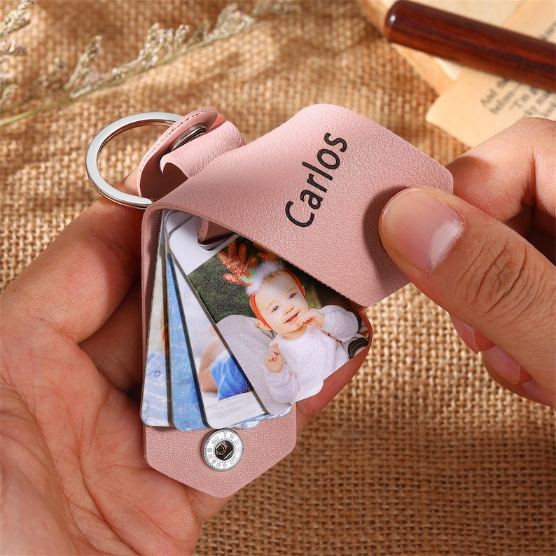 May include: A pink leather keychain with a silver ring, featuring a small photo album. The cover is embossed with the name "Carlos". The album is open, revealing several photos of a baby.