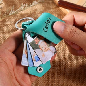 May include: A teal leather keychain with the name "Carlos" printed on it. The keychain holds several small photo cards with images of a baby. The keychain is held by a person's hand.
