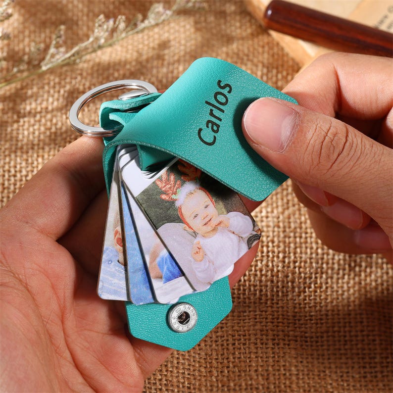 May include: A teal leather keychain with the name "Carlos" printed on it. The keychain holds several small photo cards with images of a baby. The keychain is held by a person's hand.