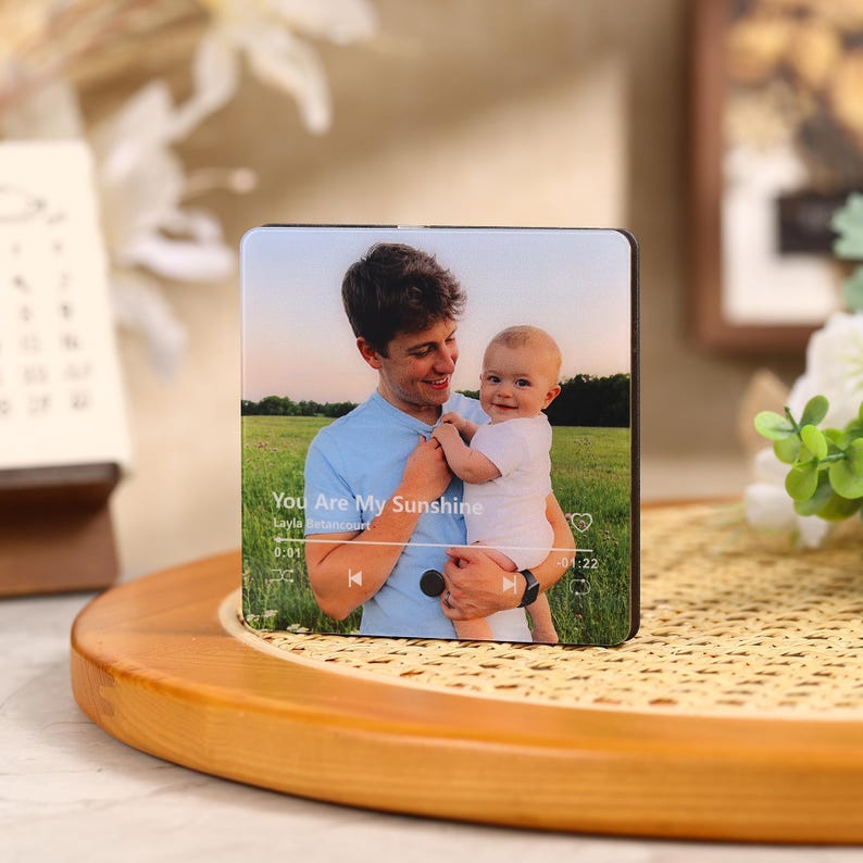 May include: A square photo block featuring a picture of a man holding a baby. The image has the text "You Are My Sunshine" and a music player interface. The block is set on a wooden tray.