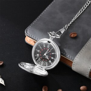 Op de afbeelding: Een zilveren zakhorloge met een zwarte wijzerplaat en Romeinse cijfers voor de uren. Het horloge heeft een ketting eraan en ligt op een grijs oppervlak.