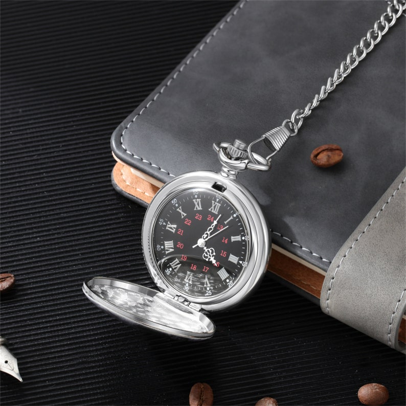 Op de afbeelding: Een zilveren zakhorloge met een zwarte wijzerplaat en Romeinse cijfers voor de uren. Het horloge heeft een ketting eraan en ligt op een grijs oppervlak.