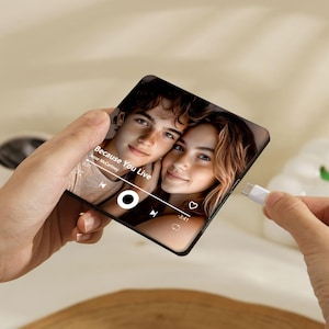 May include: A square, black phone case displaying a photo of two people with the text "Because You Live" and a music player interface. A white USB-C cable is plugged into the case.