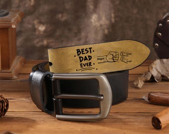 May include: A black leather belt with a silver buckle. The belt features a tan leather tag with the words "BEST DAD EVER" and illustrations of fists and names. The belt is rolled up on a wooden surface.